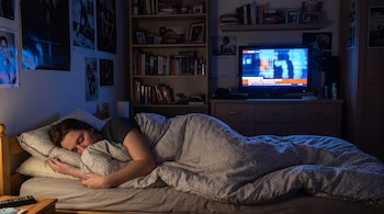 Una persona duerme en una cama con edredón y almohadas, en una habitación oscura. La televisión está encendida, mostrando imágenes. Hay un estante con libros y pósteres.