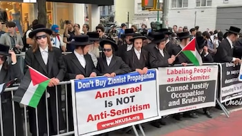 Grupo de hombres con atuendo tradicional judío ultraortodoxo sostienen banderas palestinas y pancartas antisionistas durante una protesta callejera