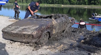 Un automóvil antiguo cubierto de barro junto a un estanque, observado por un policía y un buzo en la orilla