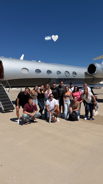 Un grupo de dieciséis personas posa delante de un avión privado gris con una escalera. En la parte superior, una bandera de Argentina y un corazón blanco