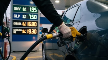 Una mano con guante blanco sostiene un surtidor amarillo introducido en el tanque de un auto junto a un letrero de precios de gasolina.