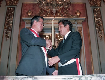 Dos hombres de traje oscuro y banda ceremonial se estrechan las manos. El hombre de la izquierda usa gafas. Detrás, una pared ornamentada con candelabro y una vela encendida