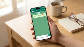 Una mano sostiene un teléfono móvil con un mensaje de 'Feliz Día del Padre' en WhatsApp. En el fondo, una mesa de madera, una taza, un cuaderno y gafas.