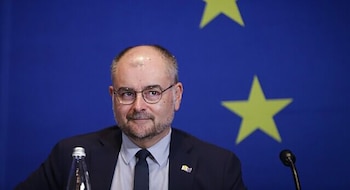 Hombre con gafas, traje y corbata frente a un fondo azul con dos estrellas amarillas de la bandera de la Unión Europea