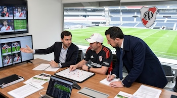 Tres hombres en una oficina, con pantalla que muestra futbolistas y formación táctica. Mesa con pizarra táctica, computadora y cuadernos. Vista a un estadio.