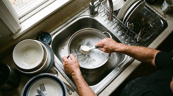 Manos de una persona lavan una olla con cepillo en un fregadero con espuma. Platos sucios apilados en encimera y cubiertos en un escurreplatos.