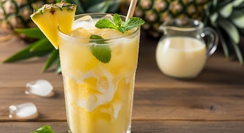 Vaso de agua de piña con leche condensada y hielo, adornado con menta y una rodaja de piña, sobre una mesa de madera con una piña entera y una jarra de leche.