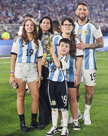 Nicolás Otamendi, su pareja Celeste Rey, y sus tres hijos, todos vestidos con camisetas de Argentina, posan en un campo de fútbol. El niño sostiene la Copa del Mundo
