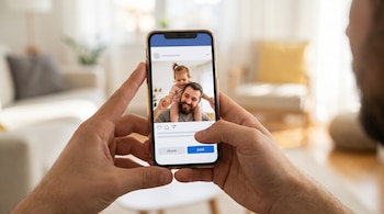 Manos de adulto sostienen un teléfono móvil con una imagen de un hombre barbudo y una niña pequeña sobre sus hombros. En la pantalla hay elementos de red social.