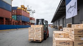 Una carretilla elevadora transporta cajas de madera con la etiqueta de salmón ahumado junto a contenedores de carga y un almacén portuario.