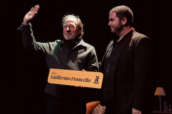 Guillermo Francella sonriente y saludando con la mano levantada, sostiene un cartel que dice 'SALA Guillermo Francella' junto a Gastón Cocchiarale en un escenario oscuro