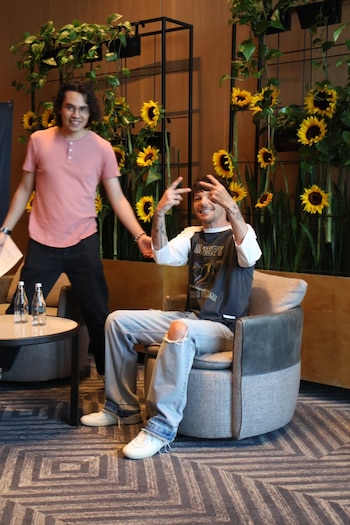 Louis Tomlinson sonríe y hace el signo de paz, sentado en un sillón gris. Un hombre de cabello rizado sonríe a su lado. Fondo con girasoles y plantas de interior