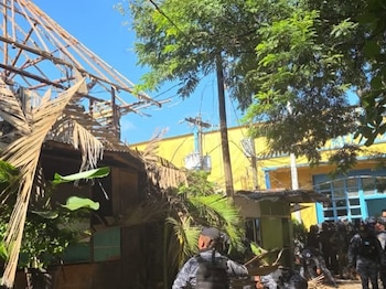 Techo de paja parcialmente destruido, vegetación verde, edificio amarillo, poste eléctrico y personas con uniformes de seguridad