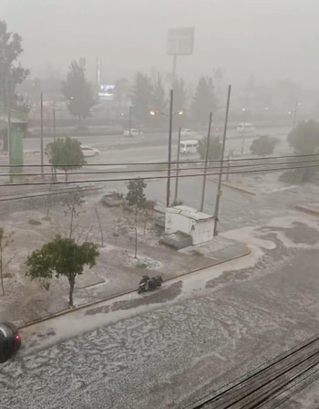 Vista aérea de una calle de la ciudad cubierta de agua y granizo durante una fuerte lluvia, con vehículos, árboles y edificios visibles a través de la neblina