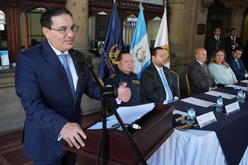 Un hombre en uniforme habla desde un podio ante una mesa con cinco personas y banderas oficiales en un salón con arcos de piedra