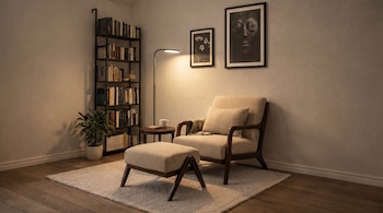 Habitación con un sillón beige, puf, y lámpara de pie en el centro. A la izquierda, una estantería con libros y una planta. Dos cuadros decoran la pared.