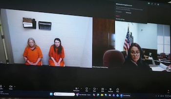 Una pantalla de ordenador muestra una videollamada judicial con dos mujeres en uniformes naranjas y una mujer en un juzgado con bandera estadounidense