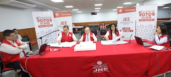 Cinco personas en chalecos rojos con el logo del JNE sentadas en una mesa roja con laptops y documentos, frente a banners de 'Recuento de Votos EG2026'