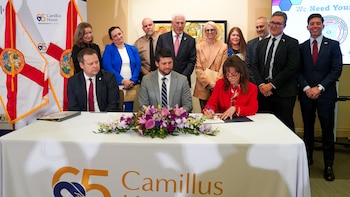 La fiscal Katherine Fernández Rundle, vestida de rojo, firma un documento en una mesa blanca con un arreglo floral. Diez personas la observan detrás, con banderas de Florida y logos de Camillus House de fondo