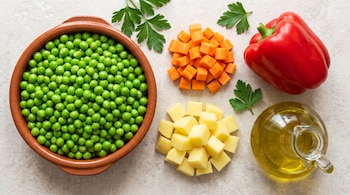 Un cuenco con arvejas, verduras cortadas en cubos, un pimiento rojo, perejil y una jarra con aceite sobre una mesa clara.