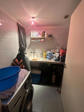 Una pequeña lavandería con paredes de azulejos blancos, un fregadero, una lavadora Samsung con un balde azul y toalla, estantes con productos de limpieza y ropa colgando