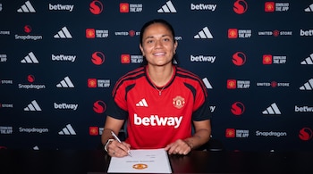 Una mujer con camiseta deportiva roja y negra firma un papel sobre una mesa negra ante una pared repleta de logotipos.