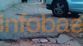 Vereda deteriorada con baldosas rotas y tierra, un tronco de árbol en el lateral y la rueda de un automóvil, con el logo de Infobae