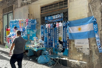 Puesto callejero con camisetas celestes, bufandas, gorras y una bandera de Argentina. Una persona camina frente al puesto en una calle de Nápoles