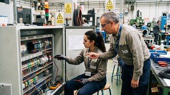 Una pasante trabaja en un panel eléctrico industrial mientras un tutor a su lado señala los componentes del tablero.