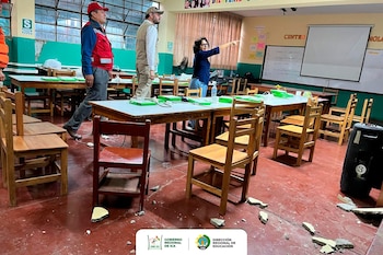 Tres personas inspeccionan un aula escolar visiblemente dañada con escombros de pared en el suelo. El mobiliario de madera está desordenado y hay grandes ventanales