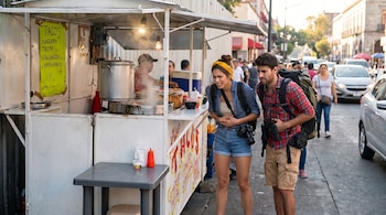 Una joven con pañuelo amarillo y un hombre con mochila, ambos con expresión de desagrado, miran un puesto de tacos con vapor en una calle urbana.