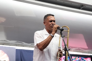 Un hombre vestido con camisa blanca canta ante un micrófono y sostiene un trombón en un escenario