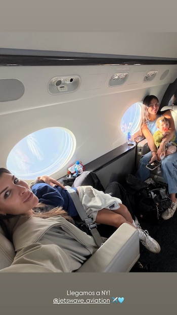 Mujer sonríe a cámara en primer plano dentro de un avión. Una mujer sostiene un bebé. Un niño duerme en el asiento. Ventanillas ovales y asientos visibles