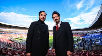 Dos hombres con vestimenta formal posan en la tribuna de un estadio de fútbol de asientos rojos y azules.