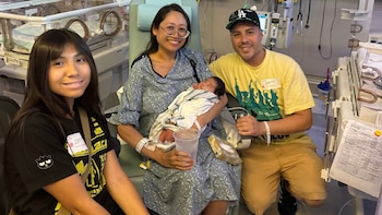 Uma família sorridente num quarto de hospital. Uma mulher de vestido azul segurando um bebê recém-nascido. Um homem e outra garota estavam ao lado dele. As incubadoras estão localizadas em segundo plano