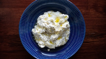 Plato redondo azul con diseño de rayas blancas contiene una porción de queso blanco grumoso con aceite de oliva y granos de pimienta negra. Fondo de madera oscura.