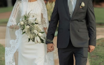 Primer plano de una pareja de novios tomados de la mano. La mujer viste de blanco con encaje y velo; el hombre, un traje oscuro con flor en la solapa