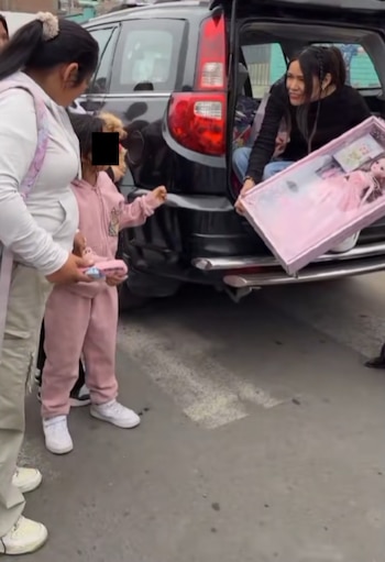 Una mujer en la cajuela de un auto negro sostiene una muñeca en caja rosa. Una niña de chándal rosa junto a una adulta extiende su brazo