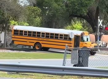 Un autobús escolar amarillo con techo blanco se encuentra atascado sobre las vías del tren junto a un paso a nivel con señalización de luz roja
