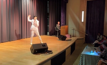 Un hombre vestido como Elvis Presley con un traje blanco y bufanda roja actúa en un escenario de madera, sosteniendo un micrófono y levantando el brazo derecho