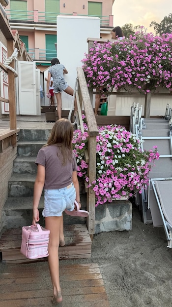 Dos niñas suben escaleras de madera con pies arenosos. Hay un bolso y sandalias rosas, jardineras con petunias rosas y blancas, y tumbonas plegables