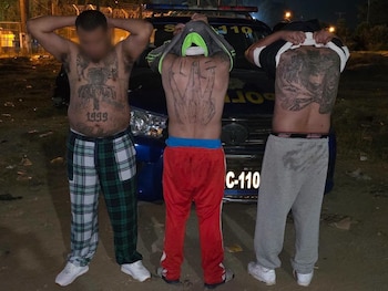 Tres hombres con torso y espalda tatuados posan de pie frente a un vehículo policial azul con luces encendidas en un entorno nocturno