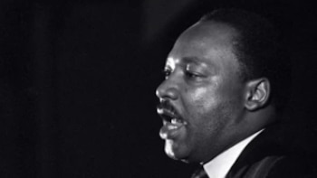 Retrato en blanco y negro de Martin Luther King Jr. hablando en un podio con micrófonos. Tiene la boca abierta, mirando hacia arriba, y viste traje y corbata