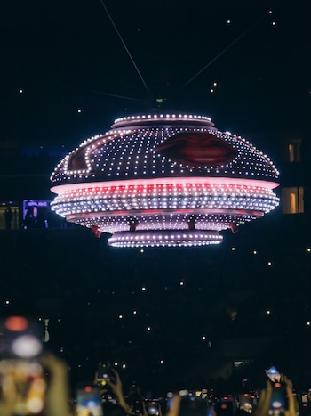 Una estructura grande con forma de disco y luces LED rojas y blancas suspendida en la oscuridad, con una multitud grabando el evento con teléfonos móviles en primer plano