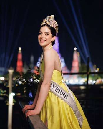 Fátima Bosch, Miss Universo, con corona y banda, sonríe a cámara en un vestido amarillo vibrante, apoyada en una barandilla de noche
