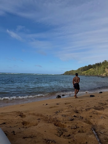 Hombre descalzo de espaldas en la orilla de una playa de arena. El mar con pequeñas olas está a su izquierda y una colina arbolada al fondo