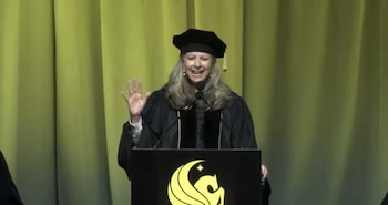 Gloria Caulfield, con toga y birrete, sonríe y levanta la mano derecha mientras habla en un podio con el logo de UCF, frente a un telón verde amarillento