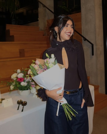 Cami Mayan sonríe sosteniendo un ramo de flores envuelto en papel blanco, vestida con top oscuro, jeans y cinturón dorado, frente a unas escaleras de madera