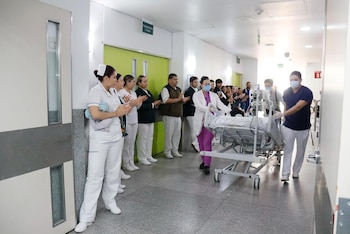 Personal médico y de enfermería aplaude a un paciente en camilla siendo trasladado por un pasillo blanco de hospital con puertas verdes y grises