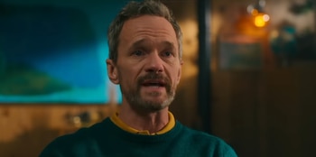 Hombre de mediana edad, pelo y barba entrecanos, vistiendo un suéter verde azulado sobre una camisa amarilla, fondo borroso con luces azules y cálidas